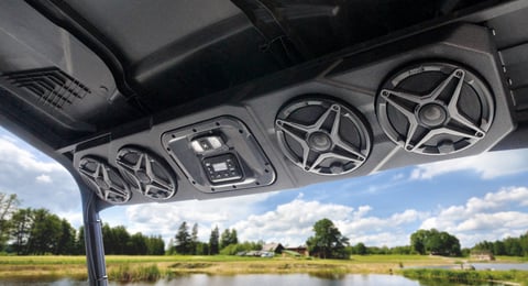 Mounted overhead speakers and control panel in a vehicle with rural landscape and water visible through the windshield
