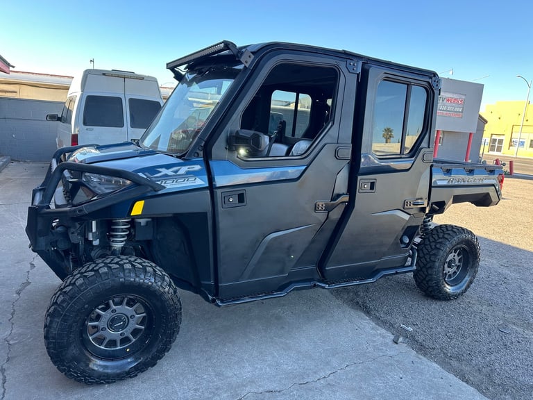 Black UTV with open hood in parking lot, showing oversized tires, extended cabin, and flatbed rear cargo area